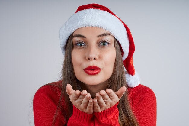 Ritratto del primo piano di bella giovane donna in maglione rosso e cappello della santa che soffia un bacio, su sfondo grigio.