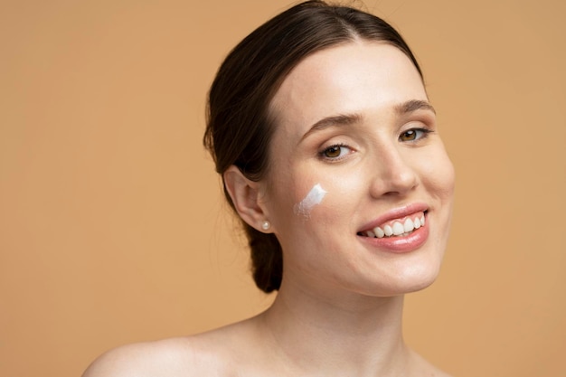 Ritratto del primo piano di bella giovane donna sorridente che applica la crema sul viso che guarda l'obbiettivo