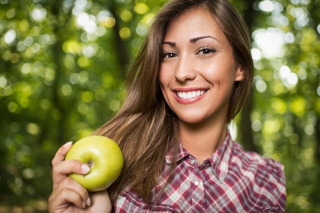 Ritratto del primo piano di bella giovane donna sorridente nella natura che tiene mela verde e che guarda l'obbiettivo