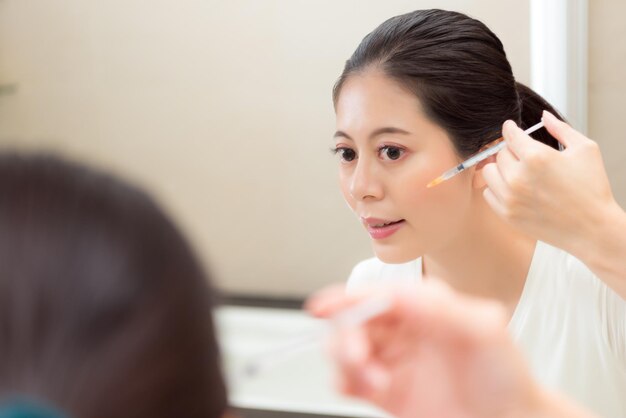 Ritratto del primo piano di giovane donna che usa il prodotto cosmetico per la cura dell'iniezione della siringa in piedi davanti allo specchio del bagno che guarda l'immagine riflessa.