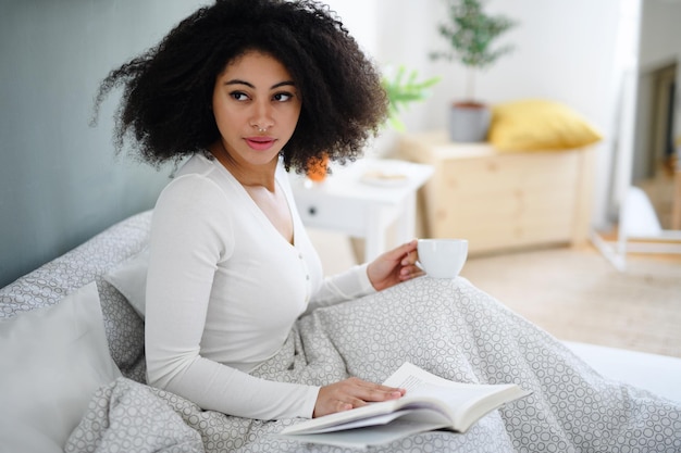 Ritratto del primo piano di giovane donna felice con libro e caffè in casa, rilassante a letto.