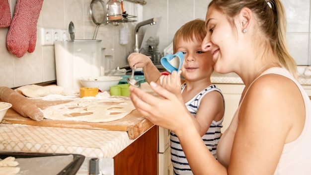 Ritratto del primo piano di giovane madre che insegna a suo figlio del bambino che fa i biscotti. Bambino con i genitori che cuociono i dolci sulla teglia in cucina in casa