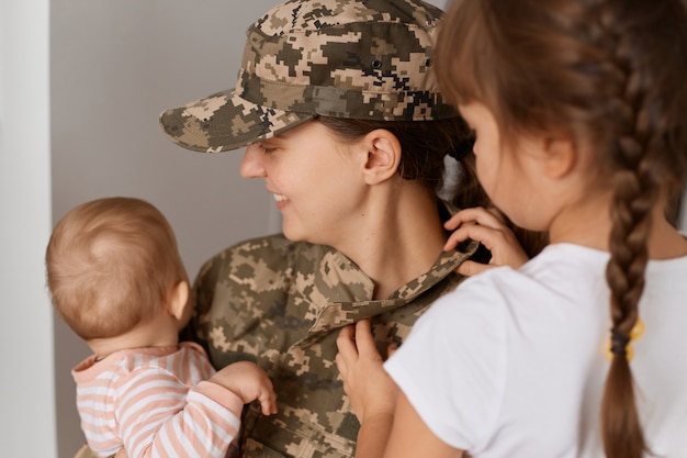 Ritratto del primo piano di giovane soldato donna adulta sorridente che indossa l'uniforme mimetica, tornando a casa dal servizio militare, abbracciando i bambini, gioendo del loro incontro.