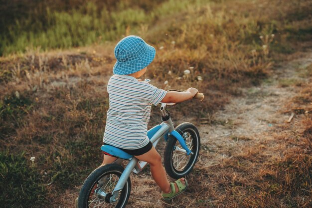 Ritratto del primo piano di un bambino che va in bicicletta su strada nella sera d'estate indossando il cappello blu vista posteriore