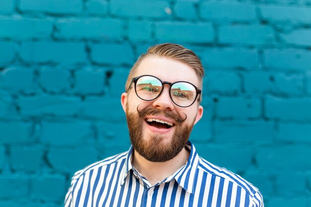 Ritratto del primo piano di un giovane sorpreso curioso in bicchieri con i baffi e la barba in posa su un muro di muro di mattoni sfocato blu. Concetto di sorpresa e informazioni scioccanti. Copyspace