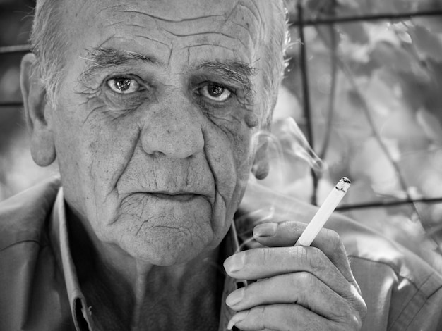 Ritratto del primo piano di un maschio pensionato greco anziano serio che fuma una sigaretta in bianco e nero
