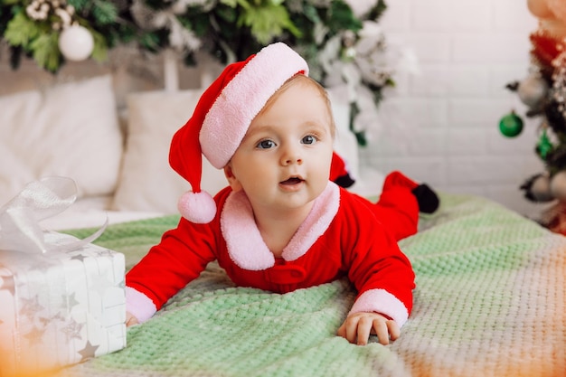 Ritratto del primo piano di un neonato. Bambino caucasico 6 mesi in un abito da Babbo Natale sdraiato su un'accogliente coperta lavorata a maglia sul letto. Natale 2022
