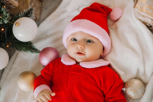 Ritratto del primo piano di un neonato. Simpatico bambino caucasico di 5-6 mesi in costume da Babbo Natale sdraiato su una coperta accogliente vicino a un albero di Natale decorato. Buon Natale 2022