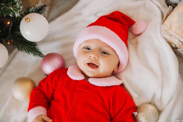 Ritratto del primo piano di un neonato. Simpatico bambino caucasico di 5-6 mesi in costume da Babbo Natale sdraiato su una coperta accogliente vicino a un albero di Natale decorato. Buon Natale 2022