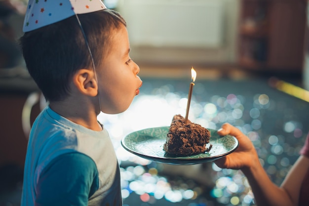 Ritratto del primo piano di un ragazzino che spegne le candeline sul pezzo di torta dato da mamma dolce foo...