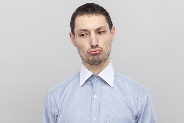 Ritratto del primo piano di un uomo d'affari di setole bello insoddisfatto triste nella classica camicia azzurra in piedi e distogliendo lo sguardo con le labbra imbronciate. studio indoor girato, isolato su sfondo grigio copyspace.