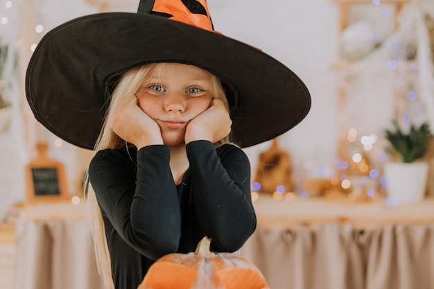 Ritratto del primo piano di una bambina bionda con gli occhi azzurri con un cappello nero di Halloween e un maglione nero