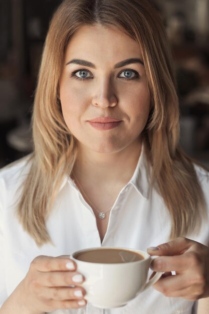 Ritratto del primo piano di una bella donna d'affari in camicia bianca dell'ufficio che beve il caffè del mattino