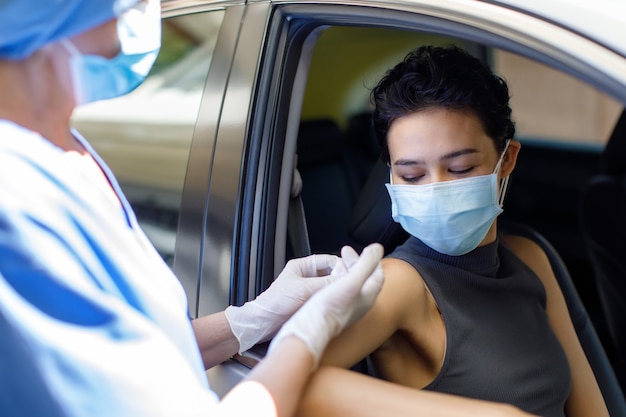 Ritratto del primo piano di una donna che indossa una maschera facciale seduta in auto che riceve il vaccino contro il coronavirus dal medico indossa l'uniforme ospedaliera usando la siringa e l'ago in guida attraverso la coda di vaccinazione.