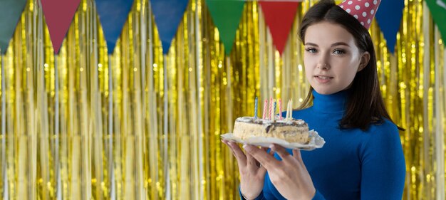 Ritratto del primo piano di una donna su sfondo giallo in un cappello da festa una bruna sta in un vestito blu con in mano una torta e si rallegra del concetto di festa di compleanno Baner