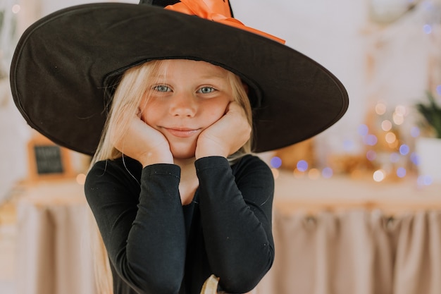 Ritratto del primo piano di una piccola ragazza bionda con gli occhi azzurri in cappello nero e maglione nero in Halloween