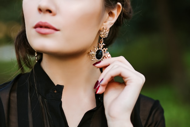 Ritratto del primo piano di una ragazza castana. Una giovane donna tocca un orecchino con pietre preziose. Orecchino in oro con pietra nera all'interno. gioielli costosi.