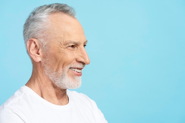 Ritratto del primo piano sorridente anziano uomo dai capelli grigi che indossa maglietta bianca isolato su sfondo blu copia spazio profilo viso attraente pensionato in posa per una foto in studio Concetto di cure dentistiche