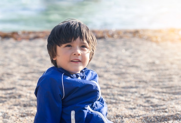 Ritratto del ragazzo felice del bambino che sorride e che cerca seduta sui ciottoli di estate di giorno soleggiato