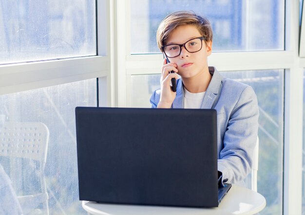 Ritratto del ragazzo teenager che parla sopra il telefono cellulare e che per mezzo del computer portatile