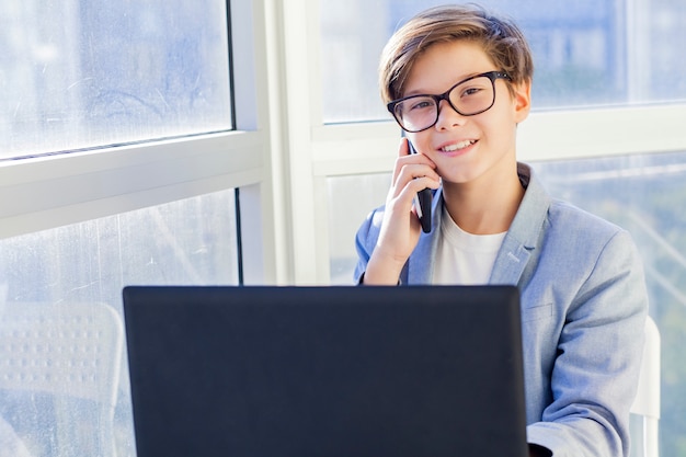 Ritratto del ragazzo teenager che parla sopra il telefono cellulare e che per mezzo del computer portatile