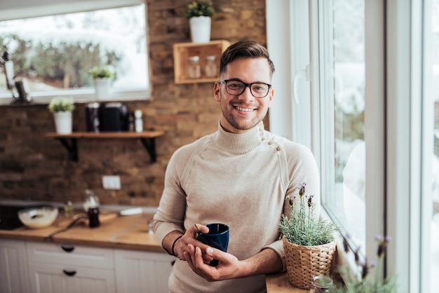 Ritratto del tè bevente sorridente dell&#39;uomo millenial vicino alla finestra a casa accogliente sulla mattina di inverno.