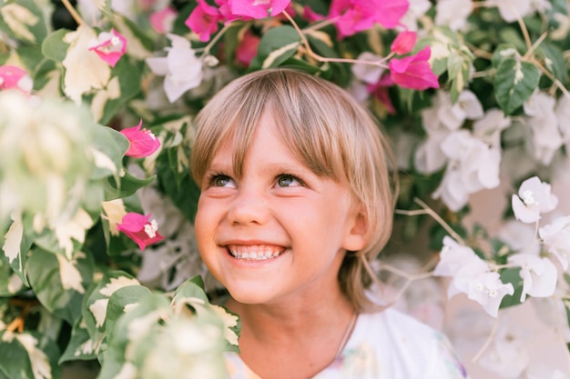 Ritratto del volto di un candido ragazzino biondo sorridente di cinque anni con gli occhi verdi in piante da fiore rosa e bianche in natura i bambini si divertono le vacanze estive luce brillante e ariosa