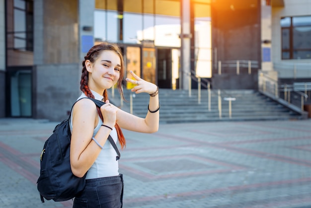 Ritratto dell'adolescente allegro attraente in maglietta bianca con lo zaino nero sulla sua spalla. Studente di liceo alla moda fa gesti con le mani