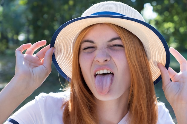 Ritratto dell'adolescente splendido in cappello giallo e con capelli rossi che mostrano la sua lingua all'aperto il giorno di estate soleggiato.