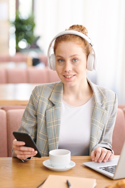 Ritratto dell'allievo adolescente bella rossa allegra in cuffie senza fili che si siede al tavolo con tazza e laptop e utilizzando l'app per smartphone nella caffetteria