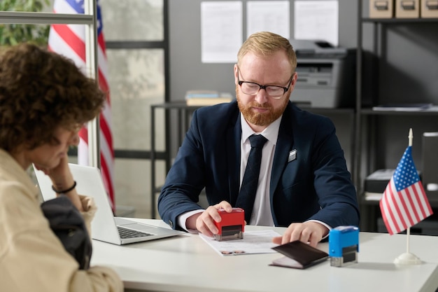 Ritratto dell'impiegato dell'immigrazione che timbra il modulo di domanda mentre lavora con il cliente nel cubicolo