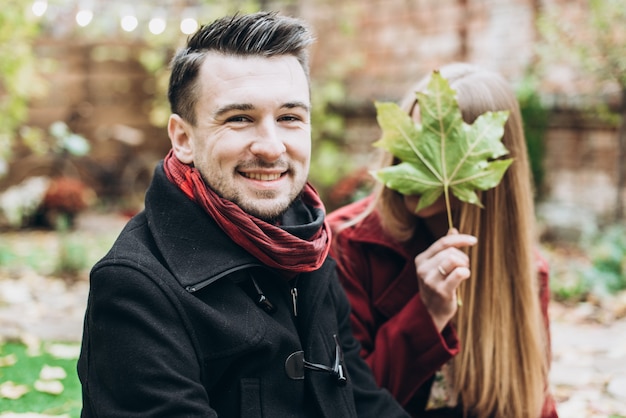 Ritratto dell'uomo allegro felice seduto sulla natura autunnale con sua moglie mentre si gode una bella giornata autunnale. Donna che si nasconde dietro la foglia d'acero