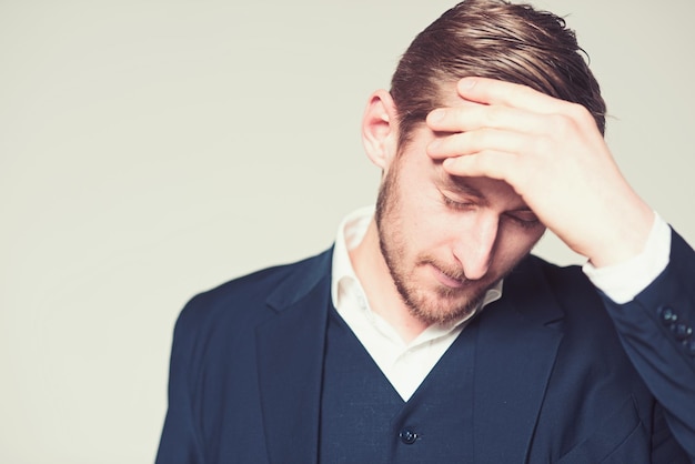 Ritratto dell'uomo d'affari pensante con la mano sulla testa Ragazzo barbuto in camicia bianca e abito blu che soffre di mal di testa Uomo biondo del primo piano isolato su sfondo grigio