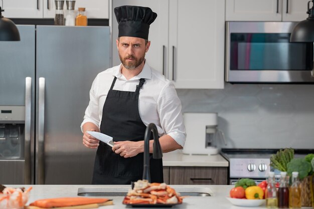 Ritratto dell'uomo in grembiule del cuoco unico e cappello del cuoco che prepara il salmone naturale fresco del pasto alla cucina bello c