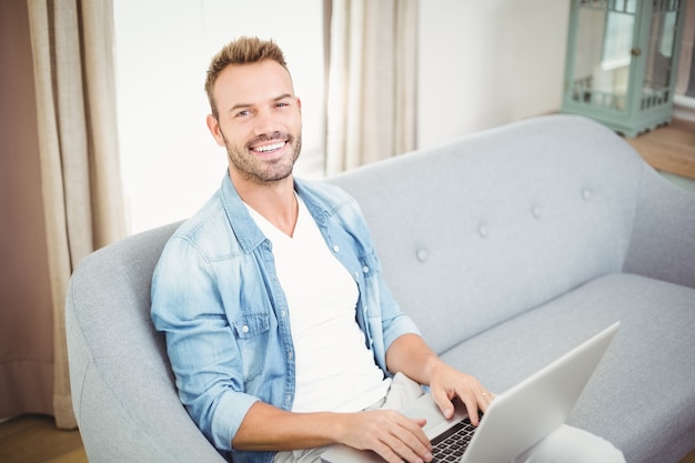 Ritratto dell'uomo sorridente che per mezzo del computer portatile a casa