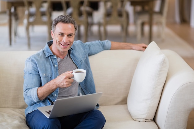 Ritratto dell'uomo sorridente che si siede sul sofà con il computer portatile che mangia caffè