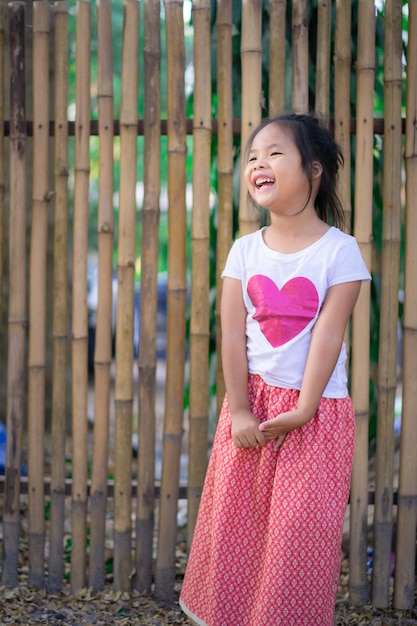 Ritratto della bambina asiatica felice che sta nel parco con la parete di bambù