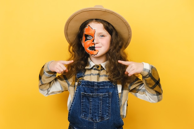 Ritratto della bambina con la maschera di trucco di Halloween che sembra fiduciosa con il sorriso sul viso, indicandosi con le dita orgogliose e felici, in posa isolato sopra la parete di fondo di colore giallo in studio