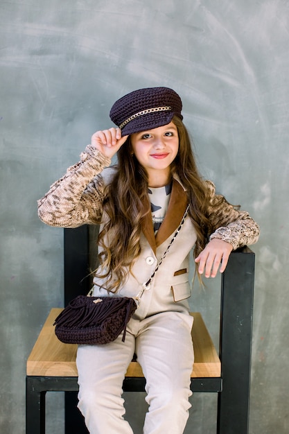 Ritratto della bambina sorridente adorabile in un vestito leggero, scarpe da tennis dorate e un cappello isolato su una seduta grigia sulla sedia