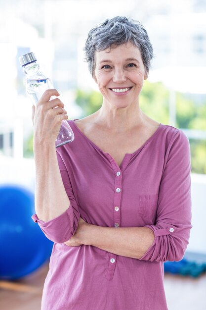 Ritratto della bottiglia di acqua matura allegra della tenuta della donna mentre stando allo studio di forma fisica