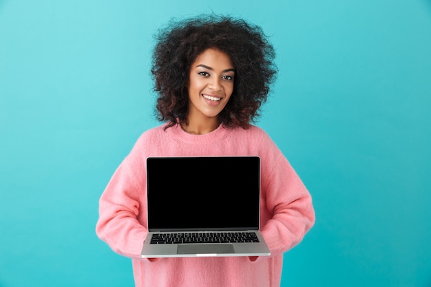 Ritratto della donna allegra 20s che tiene computer portatile e che dimostra lo schermo del nero del copyspace, isolato sopra la parete blu