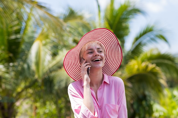 Ritratto della donna allegra in cappello rosa che fa telefonata sopra la foresta tropicale