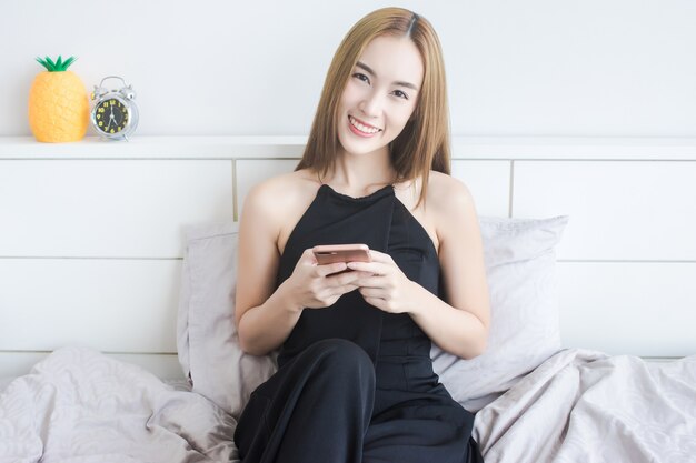 Ritratto della donna asiatica attraente che si siede sul letto mentre uso smartphone