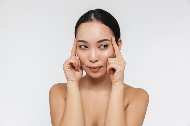 Ritratto della donna asiatica mezza nuda sorridente che tocca la sua pelle chiara e che guarda da parte isolata sopra la parete bianca