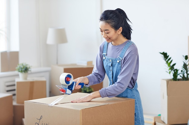 Ritratto della donna asiatica sorridente che imballa le scatole di cartone con l'erogatore del nastro mentre st si trasferisce alla nuova casa