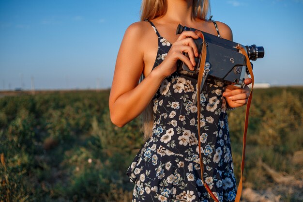 Ritratto della donna bionda in vestito dalla stampa floreale con la videocamera d'annata nel campo dell'uva durante il tramonto