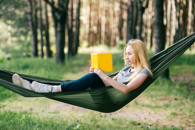 Ritratto della donna bionda preety che si rilassa in amaca in foresta soleggiata e che legge libro elettronico.