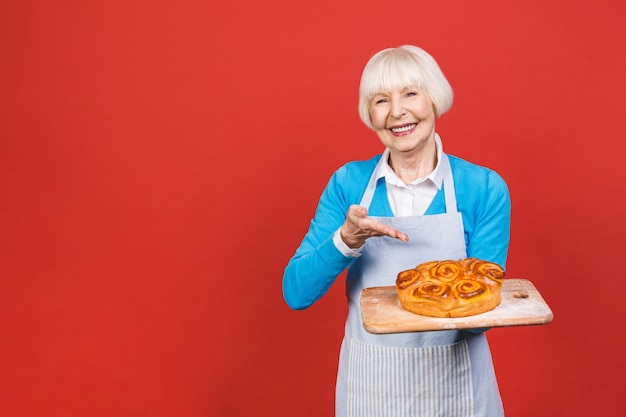 Ritratto della donna invecchiata senior allegra abbastanza affascinante con la grinza che mostra gesturing torta casalinga dolce