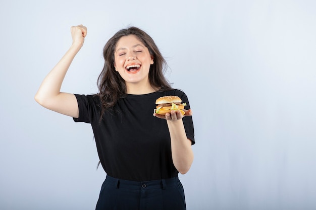 Ritratto della donna positiva che posa con l'hamburger delizioso sulla parete bianca.