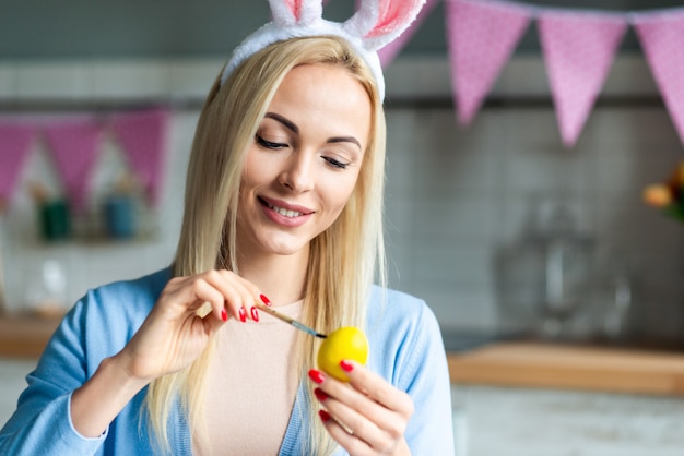 Ritratto della donna sorridente che dipinge un uovo che indossa nelle orecchie del coniglietto durante la preparazione a Pasqua.